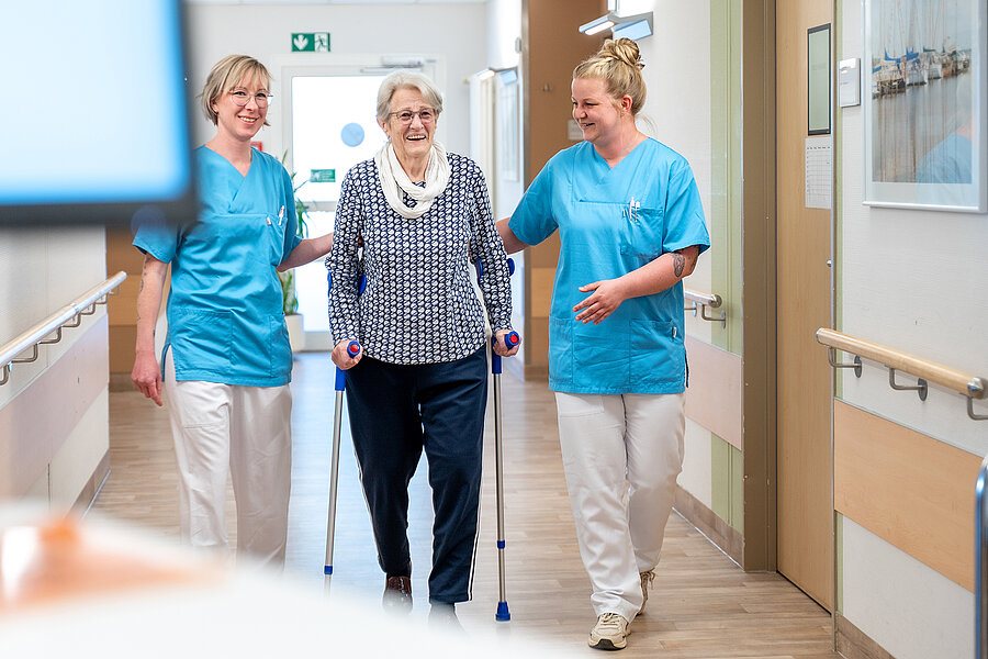 Zwei Personen in medizinischer Arbeitskleidung begleiten eine ältere Person in einem Krankenhausflur. Die ältere Person geht in der Mitte an Krücken und wird von den beiden Personen leicht gestützt.  Zwei Personen in medizinischer Arbeitskleidung begleiten eine ältere Person in einem Krankenhausflur. Die ältere Person geht in der Mitte an Krücken und wird von den beiden Personen leicht gestützt.
