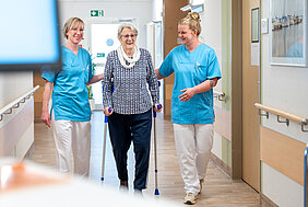 Zwei Personen in medizinischer Arbeitskleidung begleiten eine ältere Person in einem Krankenhausflur. Die ältere Person geht in der Mitte an Krücken und wird von den beiden Personen leicht gestützt.