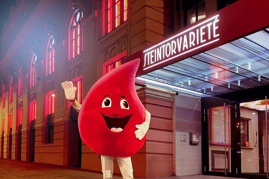 Eine Person im Maskottchen-Kostüm des Blutspendedienstes des Universitätsklinikums Halle (Saale) (ein großer Blutstropfen mit Gesicht und Armen) steht vor dem Eingang eines rot erleuchteten Hauses mit der Aufschrift Steintorvarieté.  Eine Person im Maskottchen-Kostüm des Blutspendedienstes des Universitätsklinikums Halle (Saale) (ein großer Blutstropfen mit Gesicht und Armen) steht vor dem Eingang eines rot erleuchteten Hauses mit der Aufschrift Steintorvarieté.
