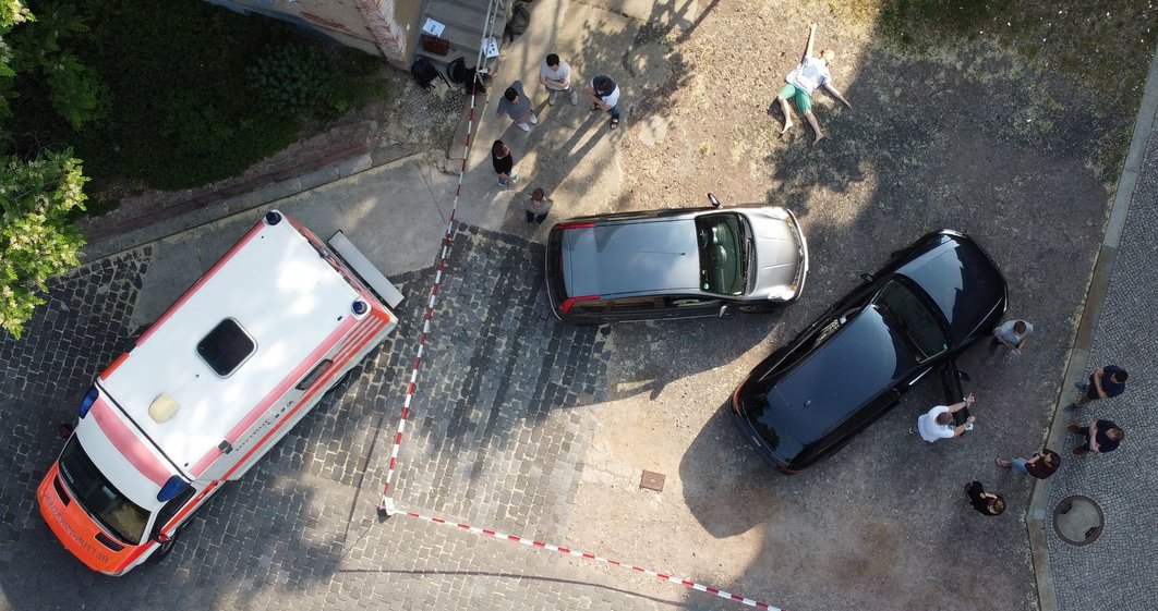 Aus der Vogelperspektive sind ein Rettungswagen, zwei Autos, eine Dummy-Puppe, die ausgestreckt auf dem Boden liegt, sowie einige stehende Personen zu sehen.