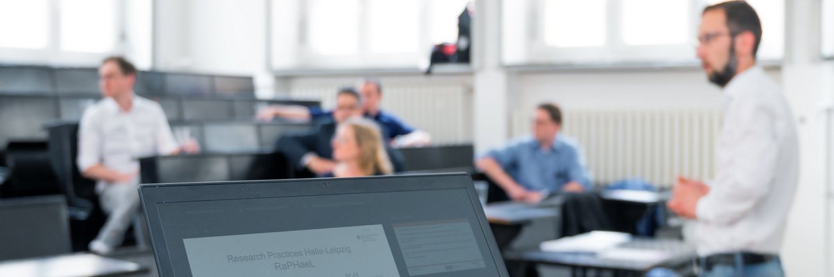 Hörsaal mit Studierenden und einem Lehrenden. Im Vordergrund ein aufgeklappter Laptop