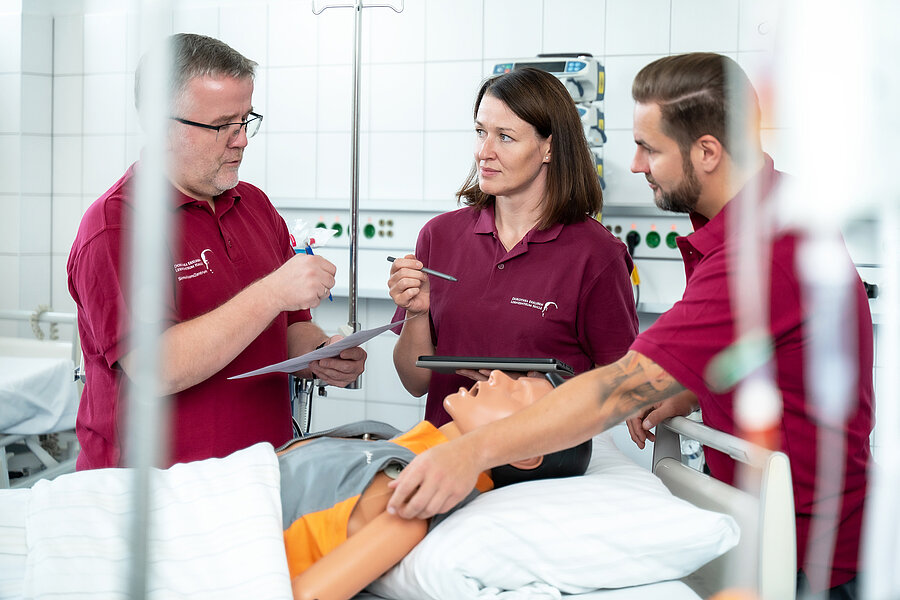 Drei Personen stehen in einem Raum des DELH. Im Vordergrund liegt eine Simulationspuppe in einem Bett. Eine Person berührt den Simulator an der Schulter, die anderen beiden Personen halten Stifte und ein Tablet bzw. ein Dokument in den Händen.  Drei Personen stehen in einem Raum des DELH. Im Vordergrund liegt eine Simulationspuppe in einem Bett. Eine Person berührt den Simulator an der Schulter, die anderen beiden Personen halten Stifte und ein Tablet bzw. ein Dokument in den Händen.