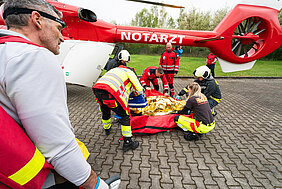 Rettungsteam versorgt verletzte Person neben einem Notarzthubschrauber auf einem Parkplatz.