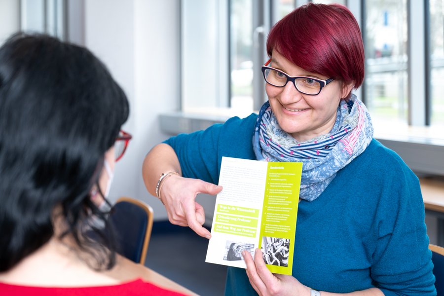 Eine Frau mit kurzen roten Haaren zeigt auf einen Infoflyer, während sie einer anderen Frau etwas beschreibt.  Eine Frau mit kurzen roten Haaren zeigt auf einen Infoflyer, während sie einer anderen Frau etwas beschreibt.