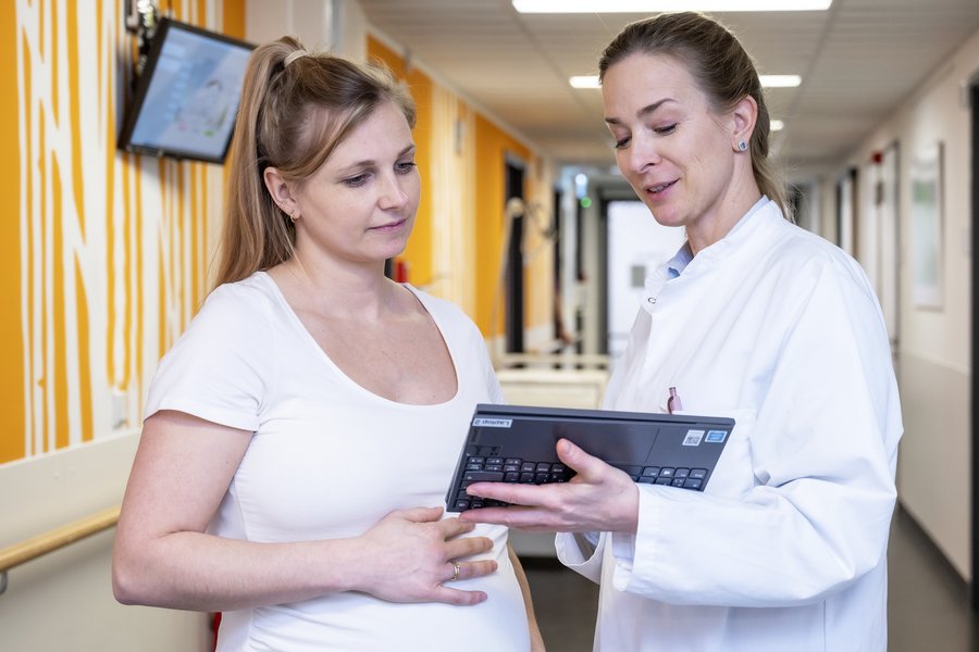 Eine Frau im weißen Kittel hält ein Tablet und ist mit einer Schwangeren mit weißem Oberteil im Gespräch. Beide stehe im Korridor einer Klinik.  Eine Frau im weißen Kittel hält ein Tablet und ist mit einer Schwangeren mit weißem Oberteil im Gespräch. Beide stehe im Korridor einer Klinik.