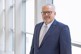 Mann mittleren Alters mit grauen Haaren, Brille, dunkler Anzug und Krawatte vor einer Fensterfront im Portrait