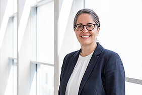 Frau mit dunklen Haaren und Brille in einem schwarzen Blazer im Portrait. Im Hintergrund ein Glasgang.