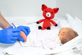 Ein kleines neugeborenes Baby liegt auf einem Wickeltisch. Das Gesicht ist nicht zu sehen. Ein Paar Hände in blauen medizinischen Einmalhandschuhen hält ein Bein des Neugeborenen fest, in der anderen Hand befindet sich eine Einmalspritze. Neben dem Neugeborenen sitzt ein gehäkelter Fuchs.