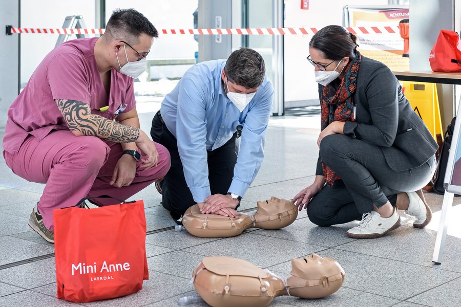 Männliche Pflegekraft in rosafarbenem Kassack hockt neben einem Mann in blauem Hemd und leitet diesen an, an einer auf dem Boden liegenden Übungspuppe eine Herzdruckmassage zu machen. Rechts hockt eine Frau in dunklem Blazer und betrachtet die Übung.  Männliche Pflegekraft in rosafarbenem Kassack hockt neben einem Mann in blauem Hemd und leitet diesen an, an einer auf dem Boden liegenden Übungspuppe eine Herzdruckmassage zu machen. Rechts hockt eine Frau in dunklem Blazer und betrachtet die Übung.