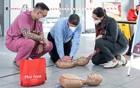 Männliche Pflegekraft in rosafarbenem Kassack hockt neben einem Mann in blauem Hemd und leitet diesen an, an einer auf dem Boden liegenden Übungspuppe eine Herzdruckmassage zu machen. Rechts hockt eine Frau in dunklem Blazer und betrachtet die Übung.