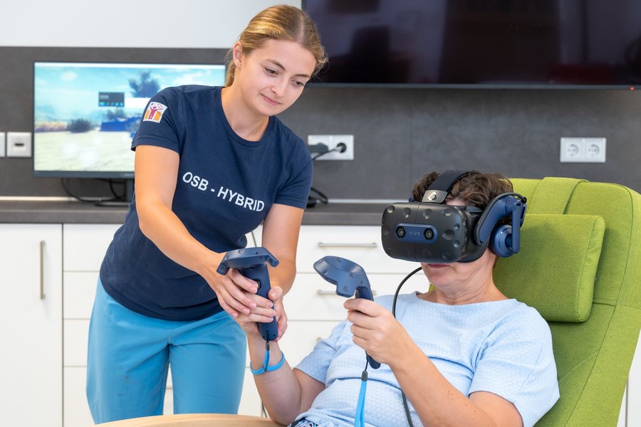 eine Frau mit VR-Brille und zwei Controllern in den Händen sitzt in einem hellgrünen Liegesessel. Links neben ihr steht eine Frau in sportlicher Arbeitskleidung und leitet sie an.  eine Frau mit VR-Brille und zwei Controllern in den Händen sitzt in einem hellgrünen Liegesessel. Links neben ihr steht eine Frau in sportlicher Arbeitskleidung und leitet sie an.