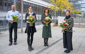 Ein Mann und drei Frauen stehen vor dem Lehrgebäude am Universitätsklinikum. Alle vier haben bunte Blumensträuße in der Hand. 