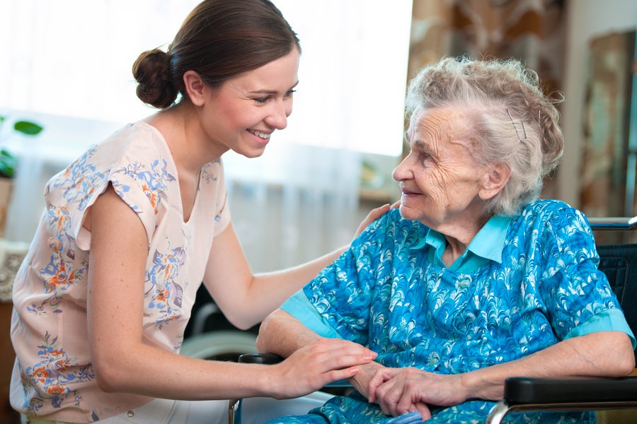 Eine junge Frau links im Bild, braune Haare hält die Hand einer Seniorin, die in einem Rollstuhl sitzt.  Eine junge Frau links im Bild, braune Haare hält die Hand einer Seniorin, die in einem Rollstuhl sitzt.