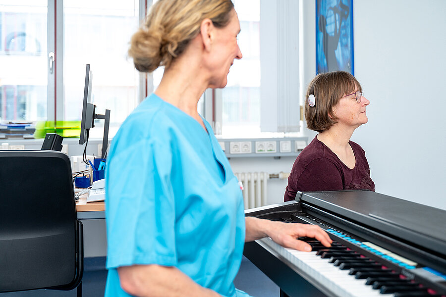 Eine Patientin sitzt mit geschlossenen Augen in einem Behandlungsraum eines Krankenhauses. An ihrem Kopf befindet sich ein Cochlea-Implantat. Im Vordergrund sitzt eine Frau in medizinischer Arbeitskleidung an einem Klavier.  Eine Patientin sitzt mit geschlossenen Augen in einem Behandlungsraum eines Krankenhauses. An ihrem Kopf befindet sich ein Cochlea-Implantat. Im Vordergrund sitzt eine Frau in medizinischer Arbeitskleidung an einem Klavier.