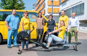 Eine Frau sitzt auf einem Ruder-Ergometer, hinter ihr stehen 3 Frauen und 3 Männer vor einem gelben Minibus. Im Bildhintergrund ist das Universitätsklinikum Halle zu sehen.