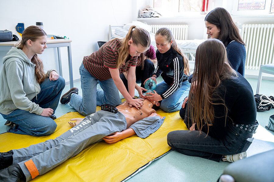 Eine Schülerin führt an einer Puppe eine Herzdruckmassage durch. Eine kleine Gruppe von Schülerinnen sitzt um die Puppe herum und schaut zu.  Eine Schülerin führt an einer Puppe eine Herzdruckmassage durch. Eine kleine Gruppe von Schülerinnen sitzt um die Puppe herum und schaut zu.