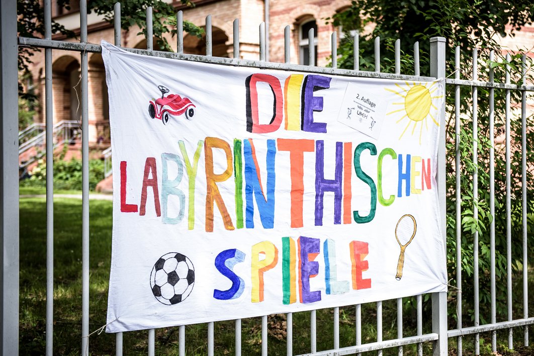 Ein Plakat aus Stoff hängt an einem Zaun. Darauf steht in bunter Schrift „Die labyrinthischen Spiele“. In die Ecken des Stoffes wurden ein kleines Fahrzeug für Kinder, eine Sonne, ein Tennisschläger und ein Fußball gemalt. Oben rechts hängt ein Zettel mit Infos zum Sportfest. 