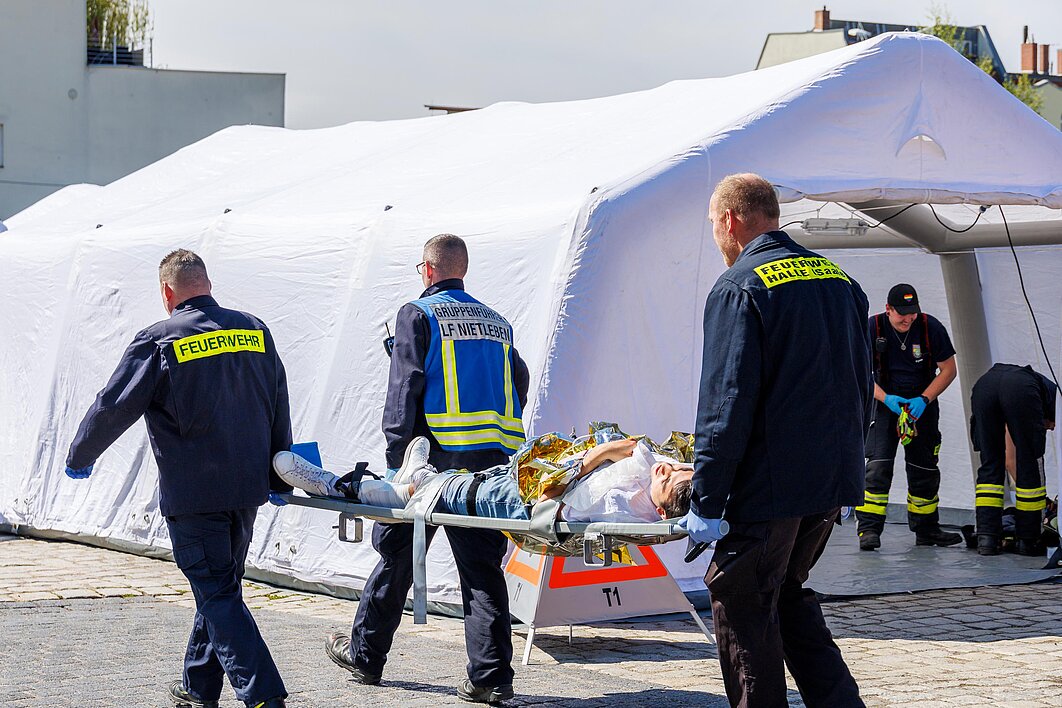 Drei Personen der Feuerwehr transportieren eine verletzte Person auf einer Trage. Im Hintergrund steht ein großes Zelt. Davor arbeiten weitere Personen.