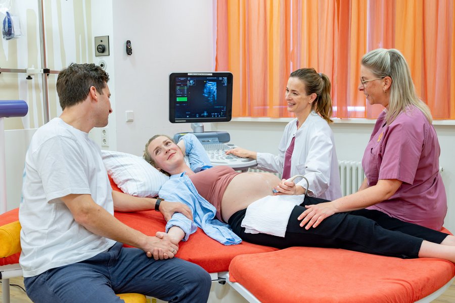 Eine Schwangere auf einer Liege wird mit Ultraschall von einer Ärztin untersucht. Eine Hebamme und ein Mann sitzen an der Liege.  Eine Schwangere auf einer Liege wird mit Ultraschall von einer Ärztin untersucht. Eine Hebamme und ein Mann sitzen an der Liege.