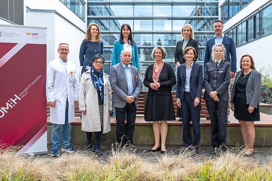 Gruppenbild von mehreren Personen auf dem Gelände des Universitätsklinikums Halle (Saale). Links im Bild steht ein Roll-up der Universitätsmedizin Halle. Die hintere Reihe der Gruppe steht etwas erhöht.  Gruppenbild von mehreren Personen auf dem Gelände des Universitätsklinikums Halle (Saale). Links im Bild steht ein Roll-up der Universitätsmedizin Halle. Die hintere Reihe der Gruppe steht etwas erhöht.