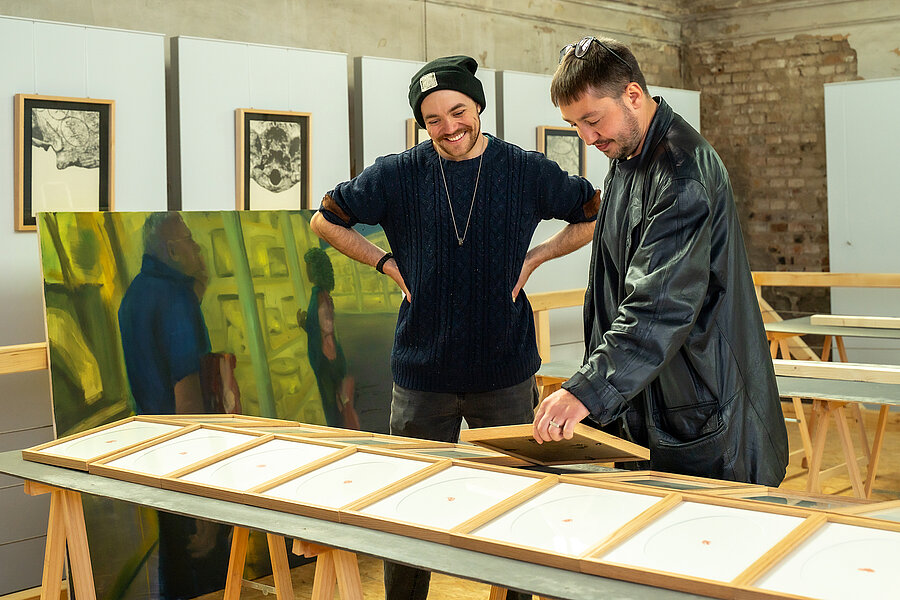 Zwei Personen stehen in einem alten Gemäuer, an dessen Wänden zahlreiche Bilderrahmen und Galeriewände lehnen. Die eine Person hat die Hände in die Hüften gestemmt und lacht, während sie ein Ausstellungsstück betrachtet. Die andere Person stellt das Exponat auf den dafür vorgesehenen Platz und lächelt.  Zwei Personen stehen in einem alten Gemäuer, an dessen Wänden zahlreiche Bilderrahmen und Galeriewände lehnen. Die eine Person hat die Hände in die Hüften gestemmt und lacht, während sie ein Ausstellungsstück betrachtet. Die andere Person stellt das Exponat auf den dafür vorgesehenen Platz und lächelt.