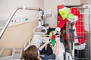 Der Grinch in einem Gitterwagen überreicht einem Jungen in einem Krankenhausbett einen mit Geschenken gefüllten Weihnachtssocken.