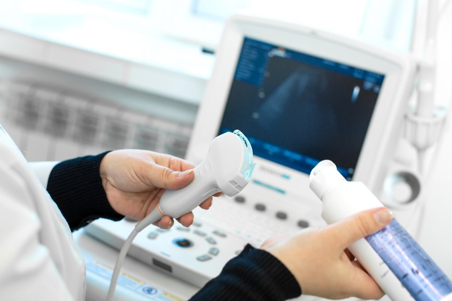 Es sind zwei Hände zu sehen, die Gleitmittel auf den Schallkopf eines Sonografie-Geräts auftragen. Im Hintergrund ist unscharf ein Ultraschallmonitor zu erkennen.   Es sind zwei Hände zu sehen, die Gleitmittel auf den Schallkopf eines Sonografie-Geräts auftragen. Im Hintergrund ist unscharf ein Ultraschallmonitor zu erkennen. 