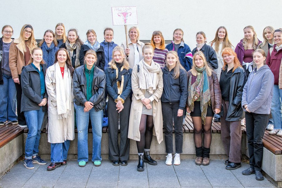 26 Personen stehen für ein Gruppenfoto in zwei Reihen vor und auf einer großen Bank und lächeln in die Kamera. In der Mitte der oberen Reihe hält eine Frau ein Schild mit der Aufschrift „HeWi‘s 2024“ und der Zeichnung einer Gebärmutter in die Höhe.  26 Personen stehen für ein Gruppenfoto in zwei Reihen vor und auf einer großen Bank und lächeln in die Kamera. In der Mitte der oberen Reihe hält eine Frau ein Schild mit der Aufschrift „HeWi‘s 2024“ und der Zeichnung einer Gebärmutter in die Höhe.