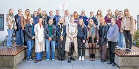 26 Personen stehen für ein Gruppenfoto in zwei Reihen vor und auf einer großen Bank und lächeln in die Kamera. In der Mitte der oberen Reihe hält eine Frau ein Schild mit der Aufschrift „HeWi‘s 2024“ und der Zeichnung einer Gebärmutter in die Höhe.