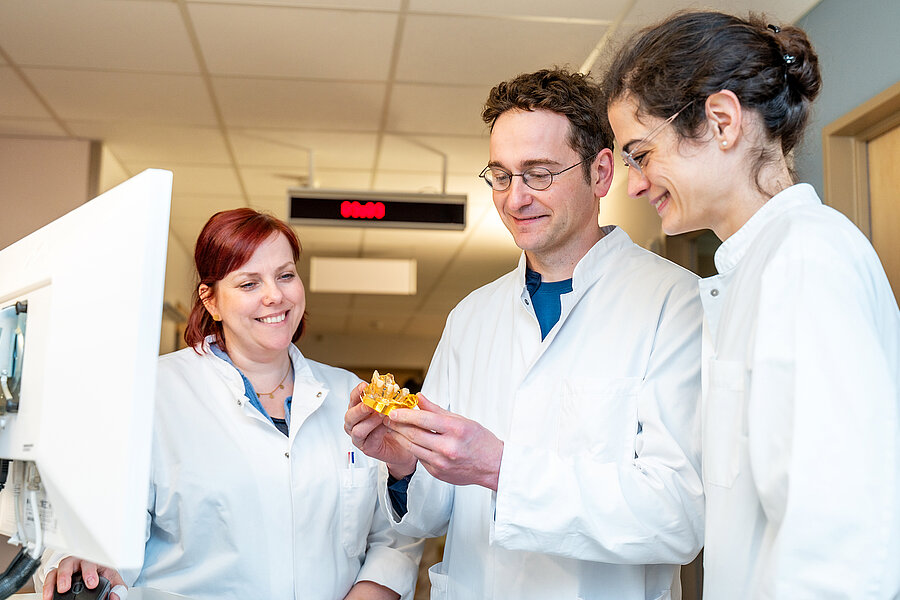 Drei Personen in medizinischer Arbeitskleidung stehen zusammen auf einem Krankenhausflur. Die Person in der Mitte hält ein kleines zahnmedizinisches Modell in der Hand.  Drei Personen in medizinischer Arbeitskleidung stehen zusammen auf einem Krankenhausflur. Die Person in der Mitte hält ein kleines zahnmedizinisches Modell in der Hand.