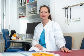 Eine Frau mit zurückgebunden brünetten Haaren sitzt mit Kittel, türkisen Kasack und Brille auf dem Kopf an einem Schreibtisch. Im Hintergrund ist ein Büro, Medizinische Ausrüstung und ein Pinguin-Kuscheltier zu erkennen.