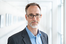 Das Bild zeigt einen Mann mittleren Alters im Anzug und blauem Hemd. Er trägt eine Brille mit kreisrunden Gläsern und einen kurz geschnittenen weißen Bart. Er hat braune, kurze Haare, blickt leicht lächelnd und souverän in die Kamera und steht in einem hell erleuchteten Korridor.