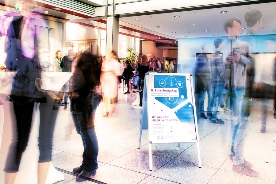 Ein mehrfach belichtetes Foto von schemenhaften menschlichen Umrissen, die in einem Foyer stehen. In der Bildmitte ist ein Posteraufsteller mit der Aufschrift "8. Forschunsgtag" zu erkennen.  Ein mehrfach belichtetes Foto von schemenhaften menschlichen Umrissen, die in einem Foyer stehen. In der Bildmitte ist ein Posteraufsteller mit der Aufschrift "8. Forschunsgtag" zu erkennen.