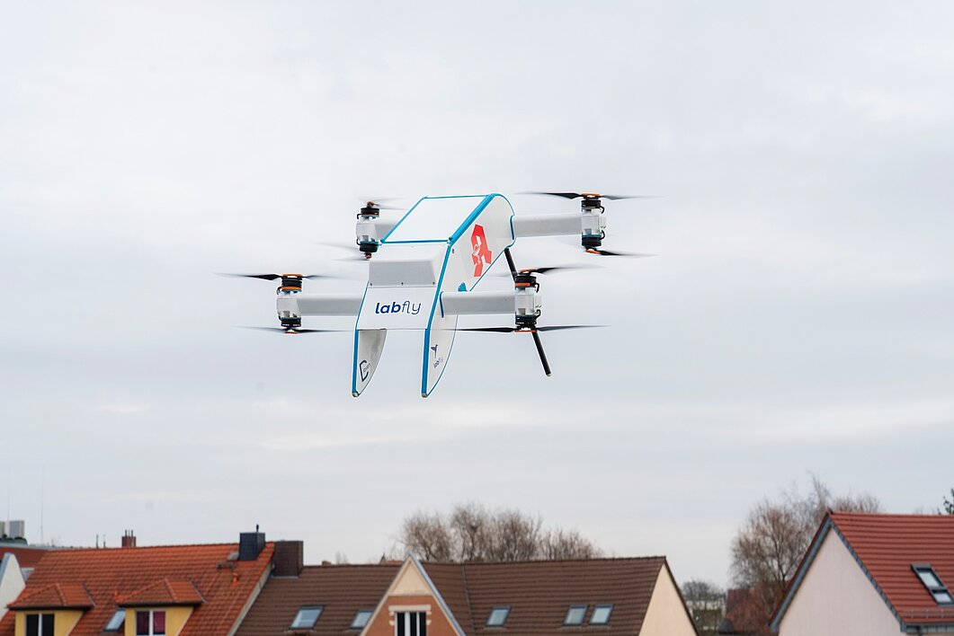 Die fliegende Drohne ist etwas größer als ein Umzugskarton, hat acht handgroße Rotoren und ist seitlich mit einem knallroten Apothekensymbol versehen. Im Hintergrund sind einige Dachspitzen von Wohnhäusern zu sehen.