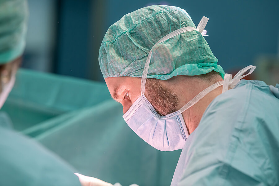 Seitliche Nahaufnahme einer Person in OP-Kleidung. Am vorderen linken Bildrand ist eine weitere Person in OP-Kleidung im Anschnitt abgebildet.  Seitliche Nahaufnahme einer Person in OP-Kleidung. Am vorderen linken Bildrand ist eine weitere Person in OP-Kleidung im Anschnitt abgebildet.