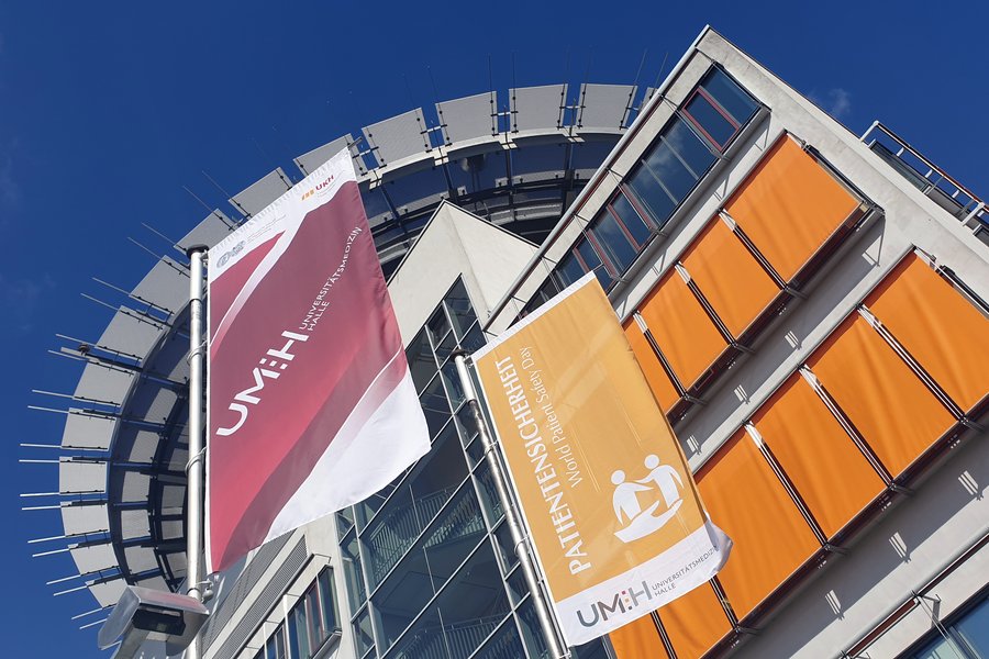 Zwei Flaggen vorm Hauptgebäude des Universitätsklinikums Halle (Saale). Die linke Fahne zeigt das Logo der Universitätsmedizin Halle auf rotem Grund, die rechte Fahne den Schriftzug "Patientensicherheit" auf Orange.  Zwei Flaggen vorm Hauptgebäude des Universitätsklinikums Halle (Saale). Die linke Fahne zeigt das Logo der Universitätsmedizin Halle auf rotem Grund, die rechte Fahne den Schriftzug "Patientensicherheit" auf Orange.