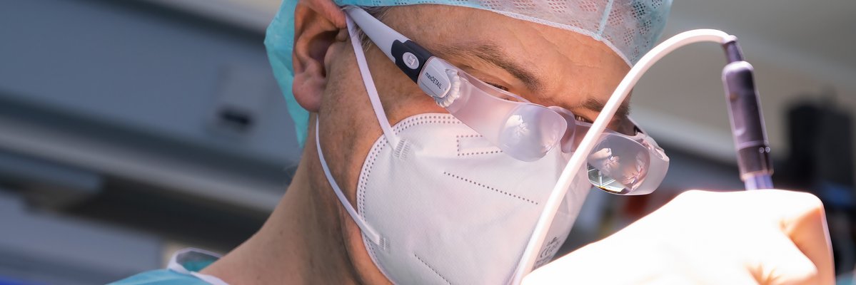 Arzt in OP-Kleidung mit Lupenbrille und einem medizinischen Gerät in der Hand