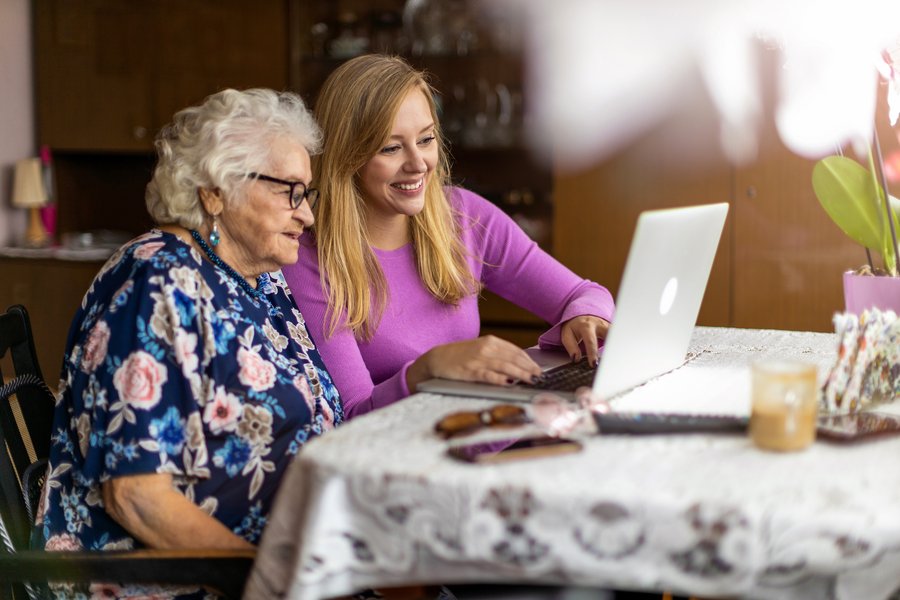 Eine ältere und eine jüngere Frau sitzen im häuslichen Umfeld vor einem Laptop an einem Tisch. Auf der Tischdecke mit Spitze liegen weitere Alltagsgegenstände wie Sonnenbrille, Handy und Fernbedienung. Die beiden Frauen lächeln und blicken auf den Laptop. Im Hintergrund ist unscharf eine große dunkelbraune Anbauwand zu erkennen.  Eine ältere und eine jüngere Frau sitzen im häuslichen Umfeld vor einem Laptop an einem Tisch. Auf der Tischdecke mit Spitze liegen weitere Alltagsgegenstände wie Sonnenbrille, Handy und Fernbedienung. Die beiden Frauen lächeln und blicken auf den Laptop. Im Hintergrund ist unscharf eine große dunkelbraune Anbauwand zu erkennen.