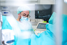 Bildvordergrund in blauer Unschärfe. Im Hintergrund ist ein Mann mit medizinischer Schutzkleidung und ein Ultraschallmonitor zu sehen.