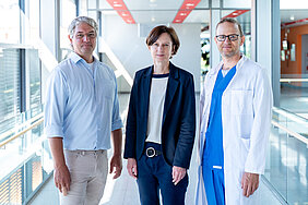 Drei Personen stehen im Gang eines Klinikums und schauen in die Kamera. Die Person rechts trägt medizinische Arbeitskleidung.