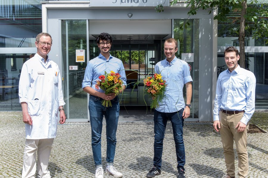 Vier Männer stehen vor dem Eingang zu einem Lehrgebäude. Zwei halten einen Blumenstrauß in der Hand  Vier Männer stehen vor dem Eingang zu einem Lehrgebäude. Zwei halten einen Blumenstrauß in der Hand