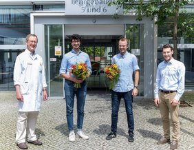 Vier Männer stehen vor dem Eingang zu einem Lehrgebäude. Zwei halten einen Blumenstrauß in der Hand