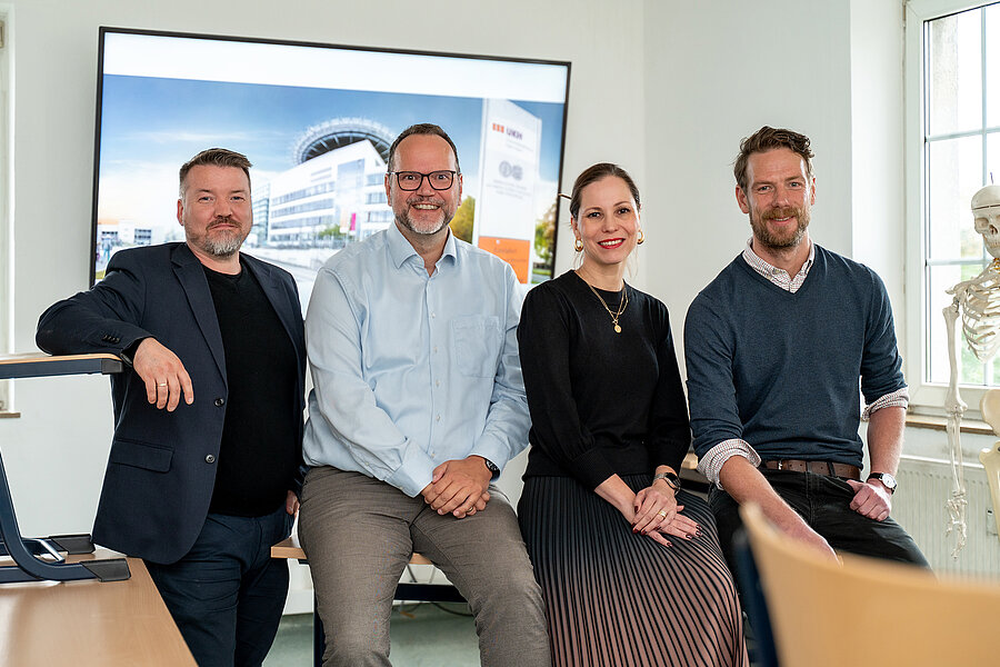 Gruppenbild mit vier Personen in einem Klassenzimmer. Drei Personen sitzen auf einem Tisch, eine vierte Person steht daneben und lehnt ihren Arm auf einen Stuhl. Im Hintergrund befindet sich ein großer Monitor mit einem Foto des Hauptgebäudes des UKH. Am rechten Bildrand befindet sich ein Skelett für den Unterricht.  Gruppenbild mit vier Personen in einem Klassenzimmer. Drei Personen sitzen auf einem Tisch, eine vierte Person steht daneben und lehnt ihren Arm auf einen Stuhl. Im Hintergrund befindet sich ein großer Monitor mit einem Foto des Hauptgebäudes des UKH. Am rechten Bildrand befindet sich ein Skelett für den Unterricht.
