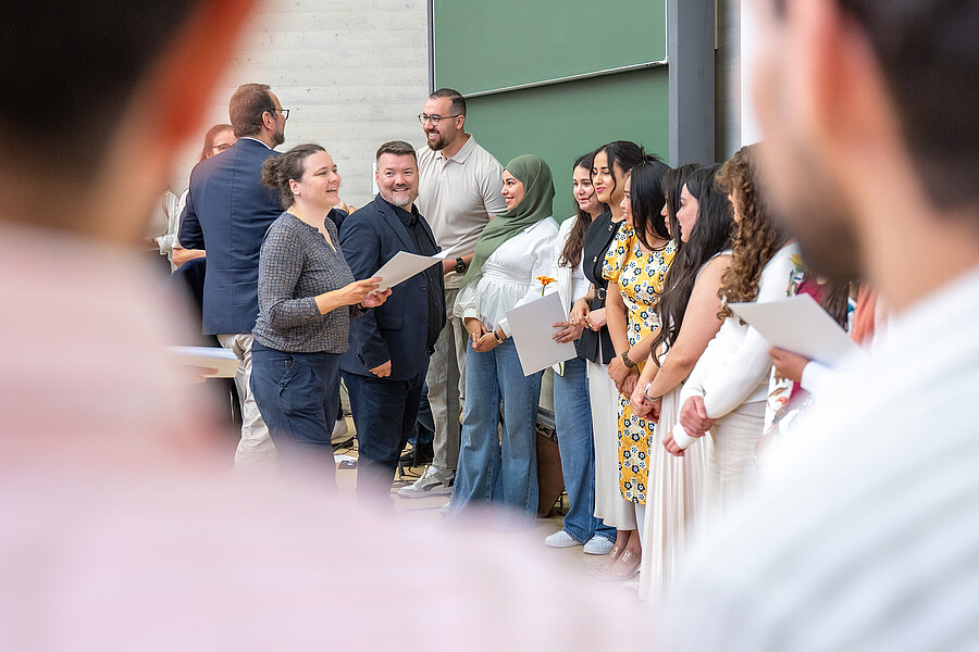 Eine Gruppe von Menschen steht in einem Hörsaal. Eine Person verteilt Abschlusszeugnisse an die Gruppe. Im Vordergrund und im Hintergrund befinden sich weitere Personen.  Eine Gruppe von Menschen steht in einem Hörsaal. Eine Person verteilt Abschlusszeugnisse an die Gruppe. Im Vordergrund und im Hintergrund befinden sich weitere Personen.