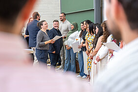 Eine Gruppe von Menschen steht in einem Hörsaal. Eine Person verteilt Abschlusszeugnisse an die Gruppe. Im Vordergrund und im Hintergrund befinden sich weitere Personen.