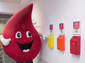 Das Maskottchen "Bob the Drop" der Blutspende des Universitätsklinikums Halle (Saale), ein großer Blutstropfen, steht neben drei Spendenboxen, die an einer Wand befestigt sind.
