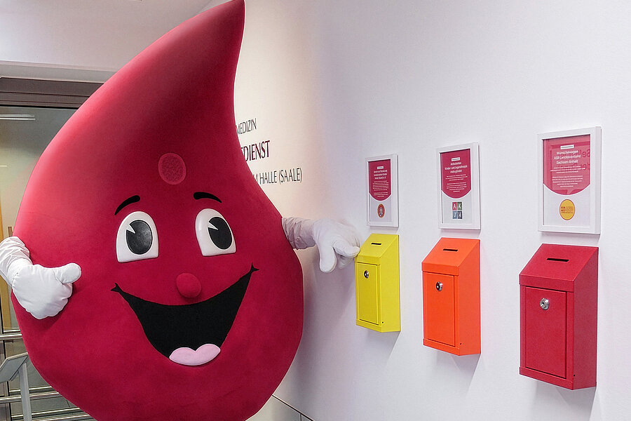 Das Maskottchen "Bob the Drop" der Blutspende des Universitätsklinikums Halle (Saale), ein großer Blutstropfen, steht neben drei Spendenboxen, die an einer Wand befestigt sind.  Das Maskottchen "Bob the Drop" der Blutspende des Universitätsklinikums Halle (Saale), ein großer Blutstropfen, steht neben drei Spendenboxen, die an einer Wand befestigt sind.
