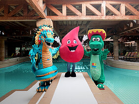 Die Maskottchen des Maya mare (links und rechts) und der Blutspende des Universitätsklinikums Halle (Saale) (Mitte) stehen zusammen am Beckenrand im Schwimmbad Maya mare. Die Maskottchen des Maya mare sind ein Drache und ein Krokodil, das Maskottchen des Klinikums ist ein großer Blutstropfen.