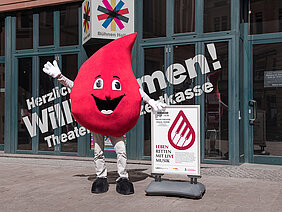 Eine Person im Maskottchen-Kostüm des Blutspendedienstes des Universitätsklinikums Halle (Saale) (ein großer Blutstropfen mit Gesicht und Armen) steht vorm Neuen Theater in Halle. Rechts neben der Person steht ein Schild mit Informationen zur Blutspendeaktion im Neuen Theater. Im Hintergrund an der Fensterfront steht ein Schriftzug. Darüber befindet sich das Logo der Bühnen Halle.