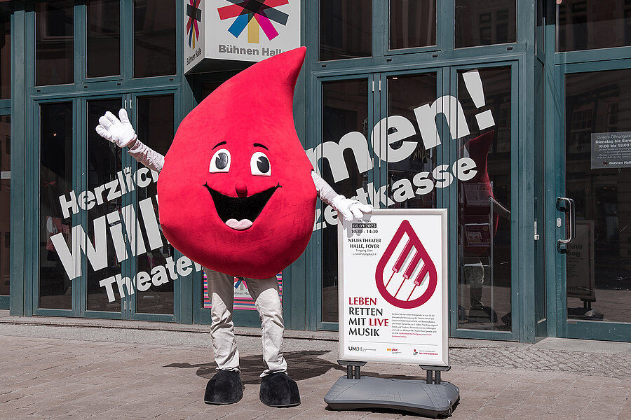Eine Person im Maskottchen-Kostüm des Blutspendedienstes des Universitätsklinikums Halle (Saale) (ein großer Blutstropfen mit Gesicht und Armen) steht vorm Neuen Theater in Halle. Rechts neben der Person steht ein Schild mit Informationen zur Blutspendeaktion im Neuen Theater. Im Hintergrund an der Fensterfront steht ein Schriftzug. Darüber befindet sich das Logo der Bühnen Halle.  Eine Person im Maskottchen-Kostüm des Blutspendedienstes des Universitätsklinikums Halle (Saale) (ein großer Blutstropfen mit Gesicht und Armen) steht vorm Neuen Theater in Halle. Rechts neben der Person steht ein Schild mit Informationen zur Blutspendeaktion im Neuen Theater. Im Hintergrund an der Fensterfront steht ein Schriftzug. Darüber befindet sich das Logo der Bühnen Halle.